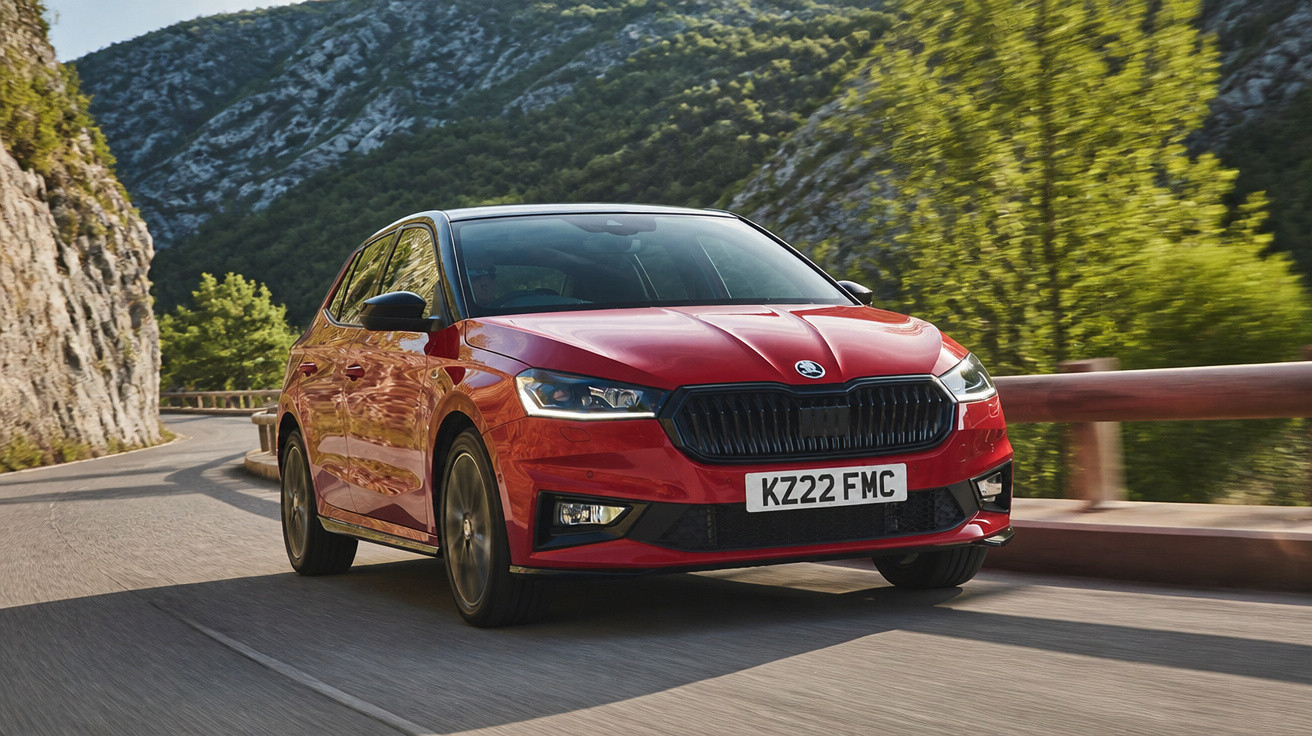 Carro vermelho Škoda a circular numa estrada de montanha com vegetação e rochas ao fundo.