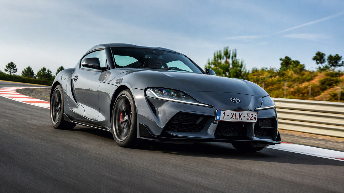 Carro desportivo cinzento Toyota Supra em curva na pista de corrida com árvores ao fundo.
