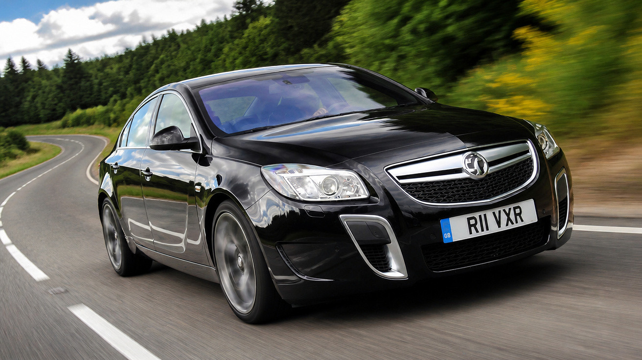 Carro preto em movimento numa estrada sinuosa rodeada de árvores verdes e céu nublado.