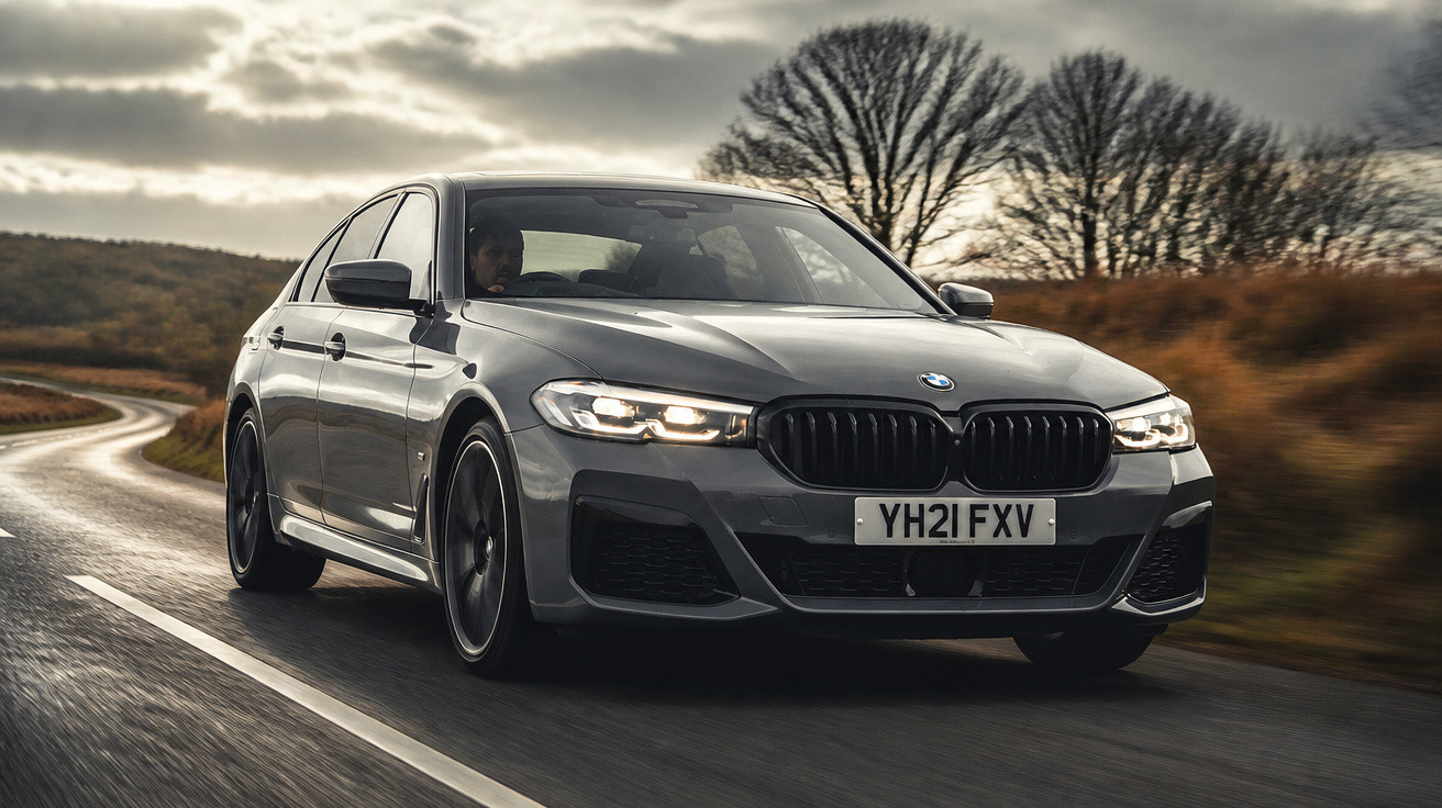 Carro BMW cinzento a conduzir numa estrada sinuosa em ambiente rural com céu nublado e árvores sem folhas.