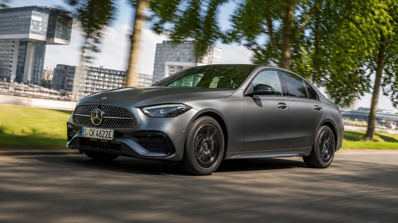 Carro Mercedes-Benz cinzento a circular numa estrada urbana com árvores e edifícios ao fundo.