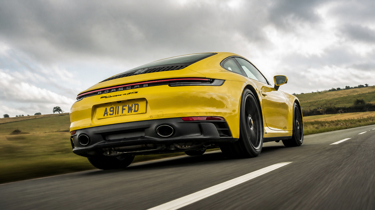 Carro desportivo amarelo Porsche 911 a circular numa estrada sob céu nublado.