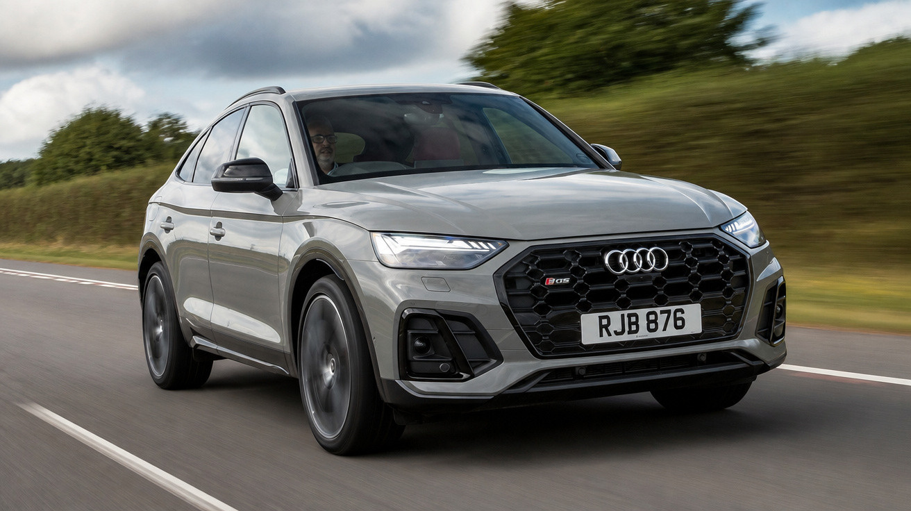 Automóvel Audi Q5 cinzento a circular numa estrada com vegetação e céu nublado de fundo.