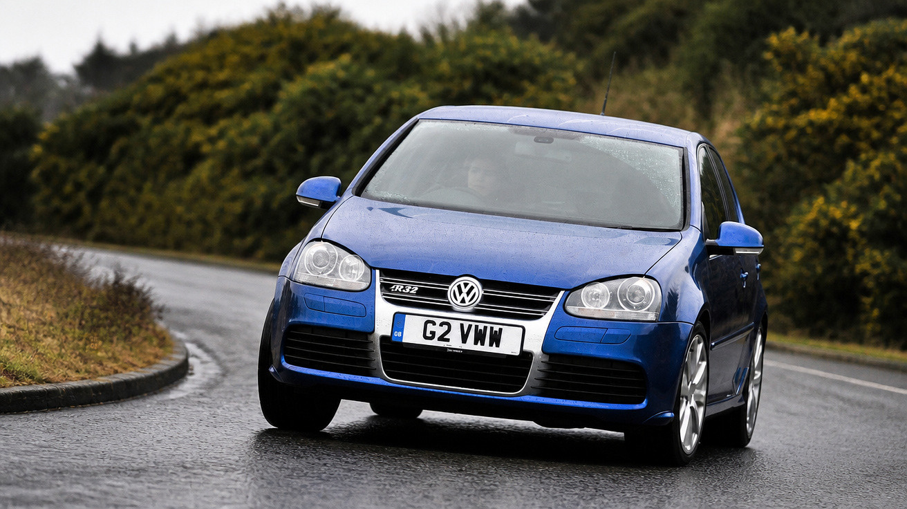 Carro azul Volkswagen Golf R32 a circular numa estrada curva e molhada com vegetação ao fundo.