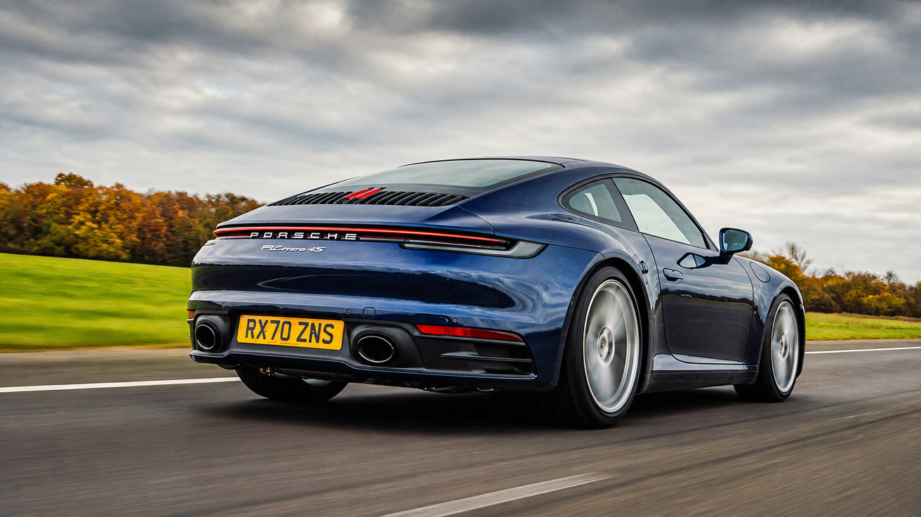Carro desportivo Porsche 911 Carrera 4S azul em movimento numa estrada com céu nublado e campo verde ao lado.