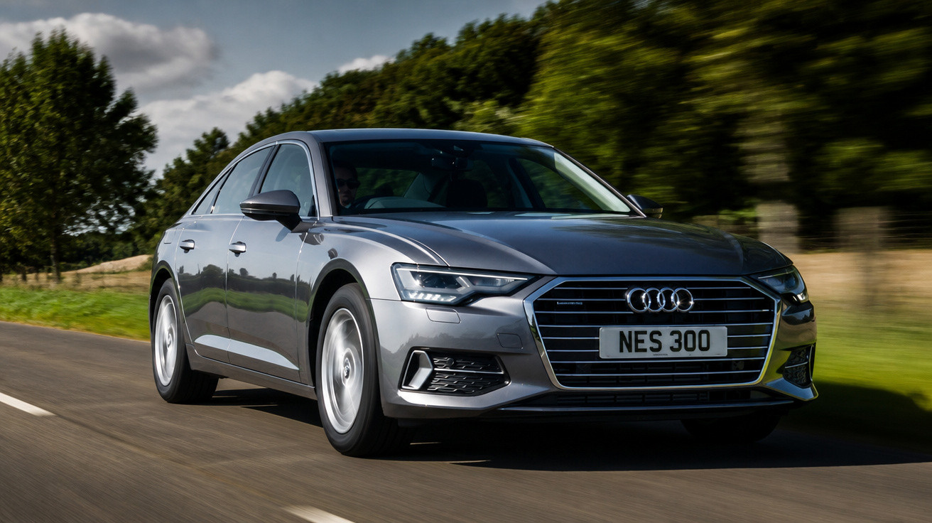 Carro cinzento Audi a circular numa estrada rodeada de árvores e relva verde.