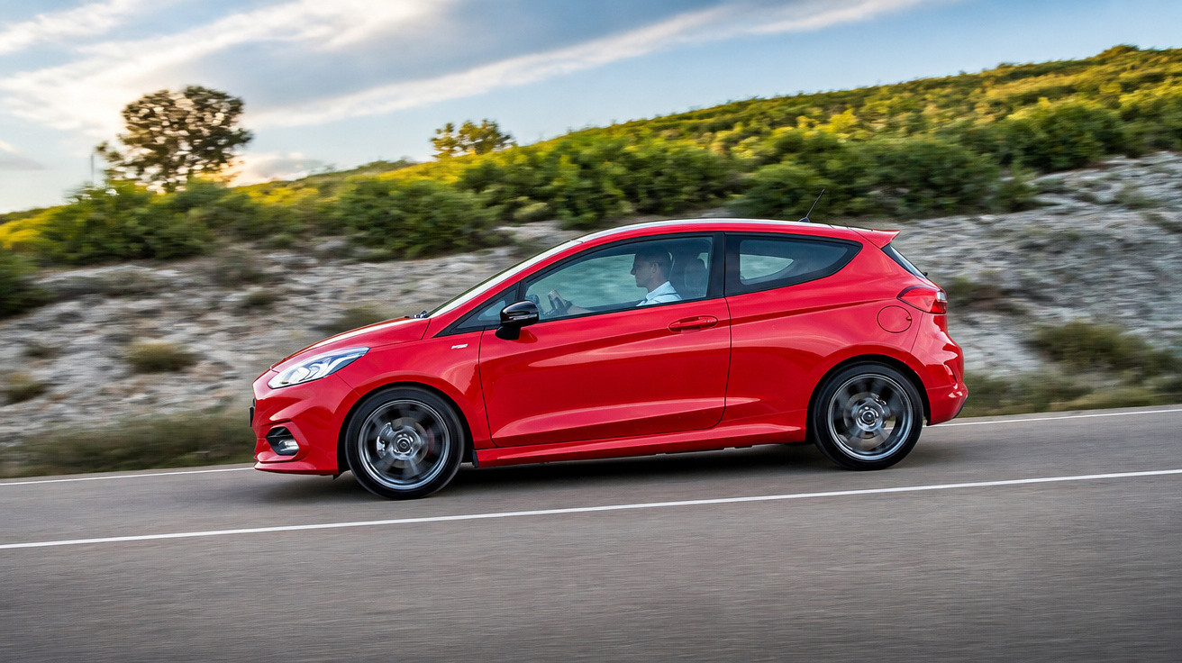 Carro vermelho desportivo a circular numa estrada asfaltada com vegetação e céu nublado ao fundo.