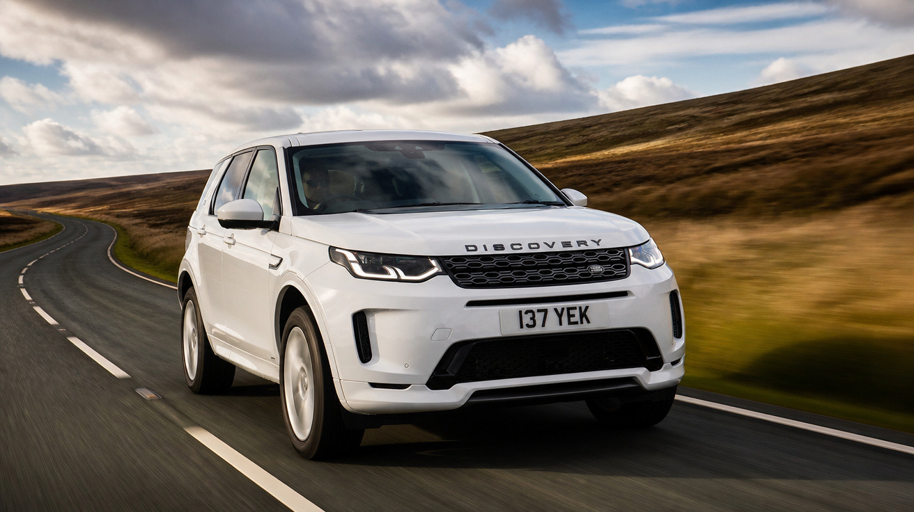 Carro Land Rover Discovery branco a circular numa estrada sinuosa rodeada por colinas e céu nublado.