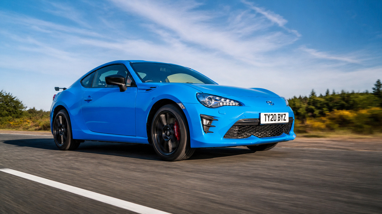 Carro desportivo azul em movimento numa estrada reta com céu azul e vegetação ao fundo.