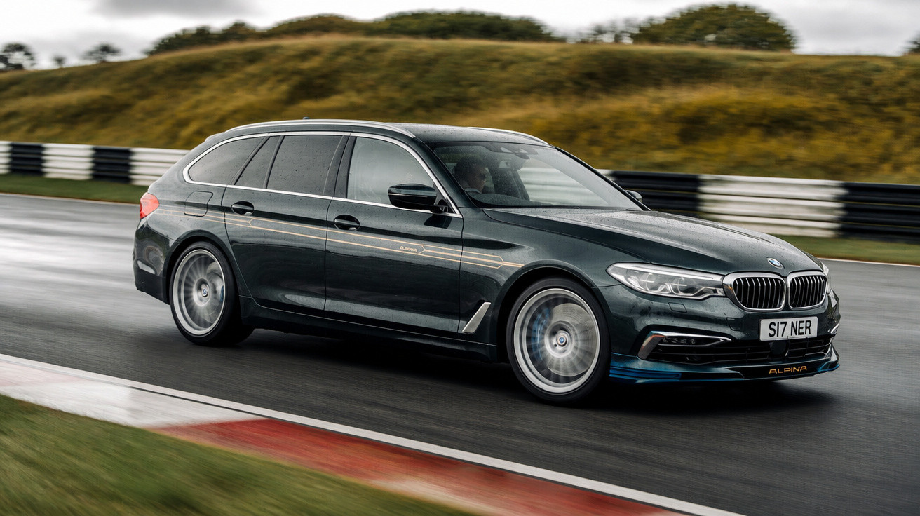 Carro BMW Alpina Touring Station Wagon a alta velocidade numa pista molhada com relva e barreiras ao fundo.