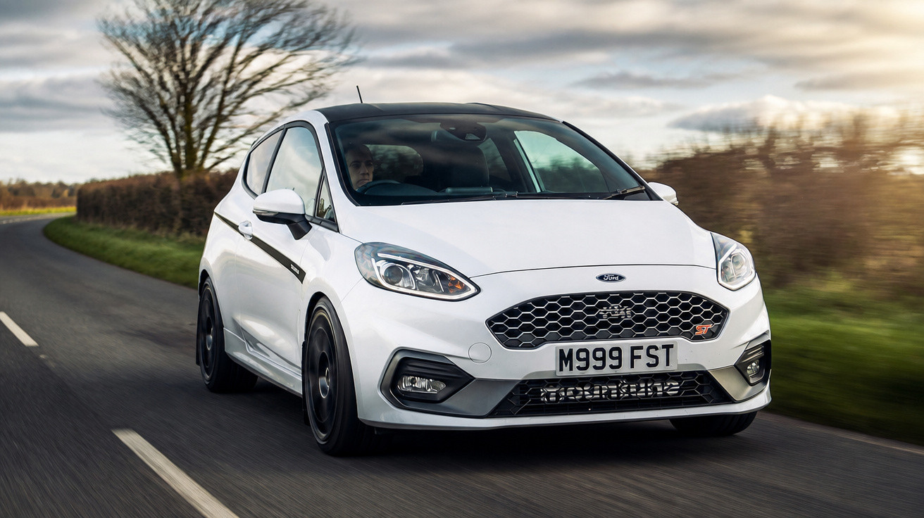Carro compacto branco Ford Fiesta ST a circular numa estrada rural com vegetação e árvores ao fundo.