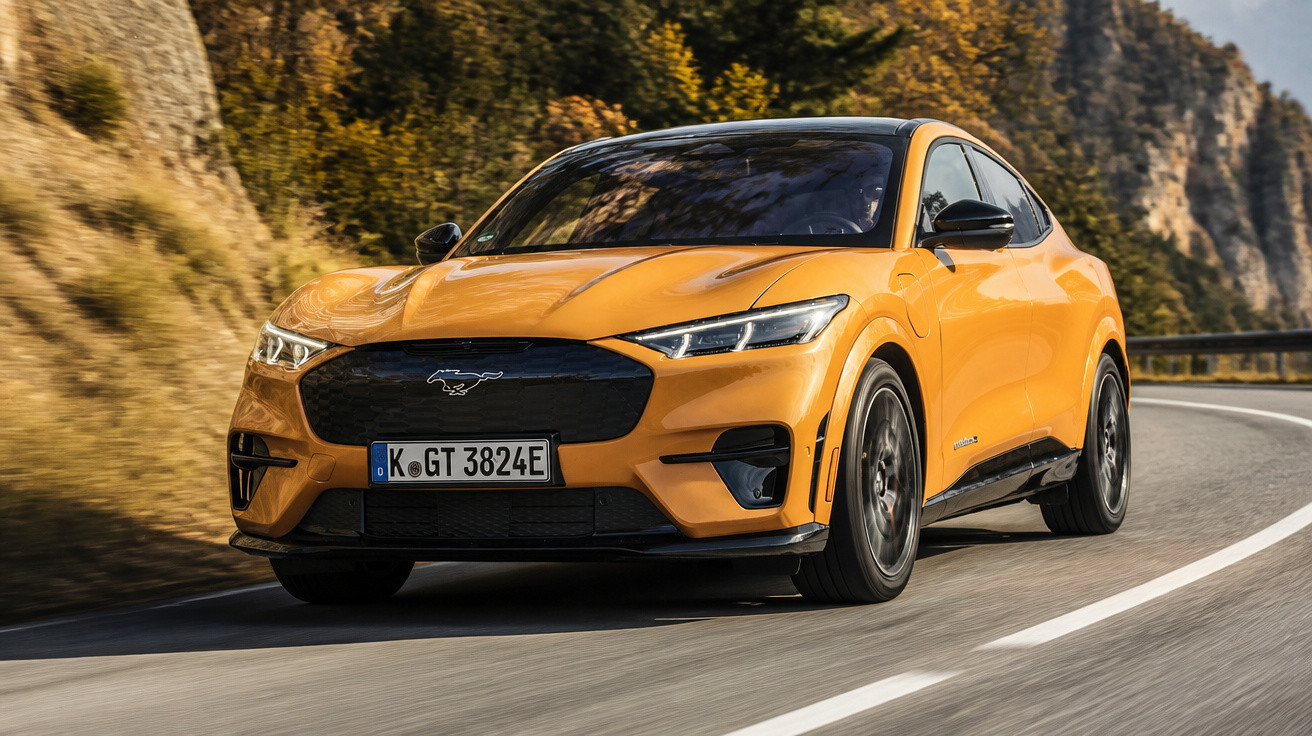 Carro desportivo amarelo Ford Mustang Mach-E a conduzir numa estrada sinuosa rodeada de árvores e montanhas.