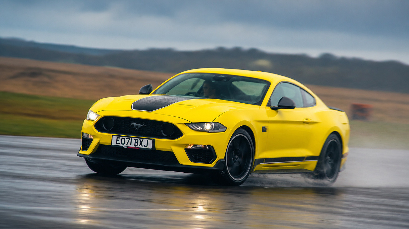 Carro desportivo amarelo a derrapar numa estrada molhada com paisagem montanhosa ao fundo.
