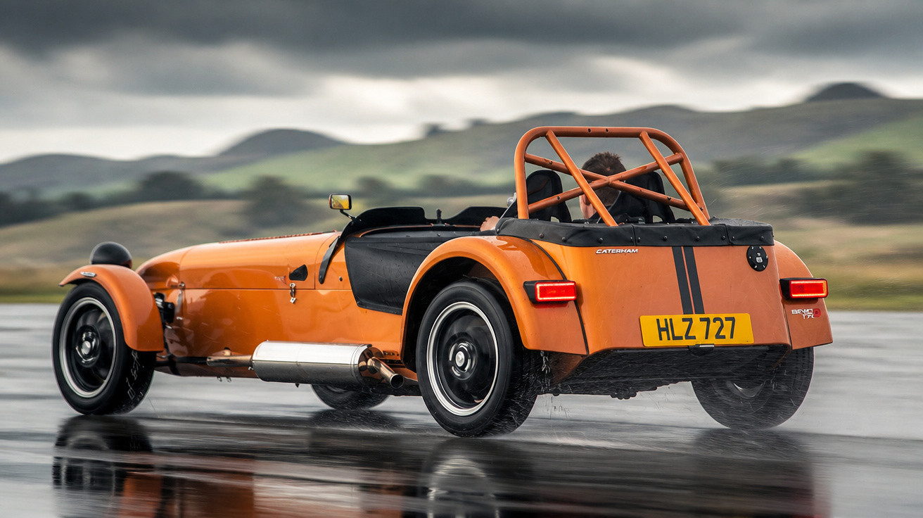 Carro desportivo laranja Caterham com dois ocupantes em estrada molhada e céu nublado.