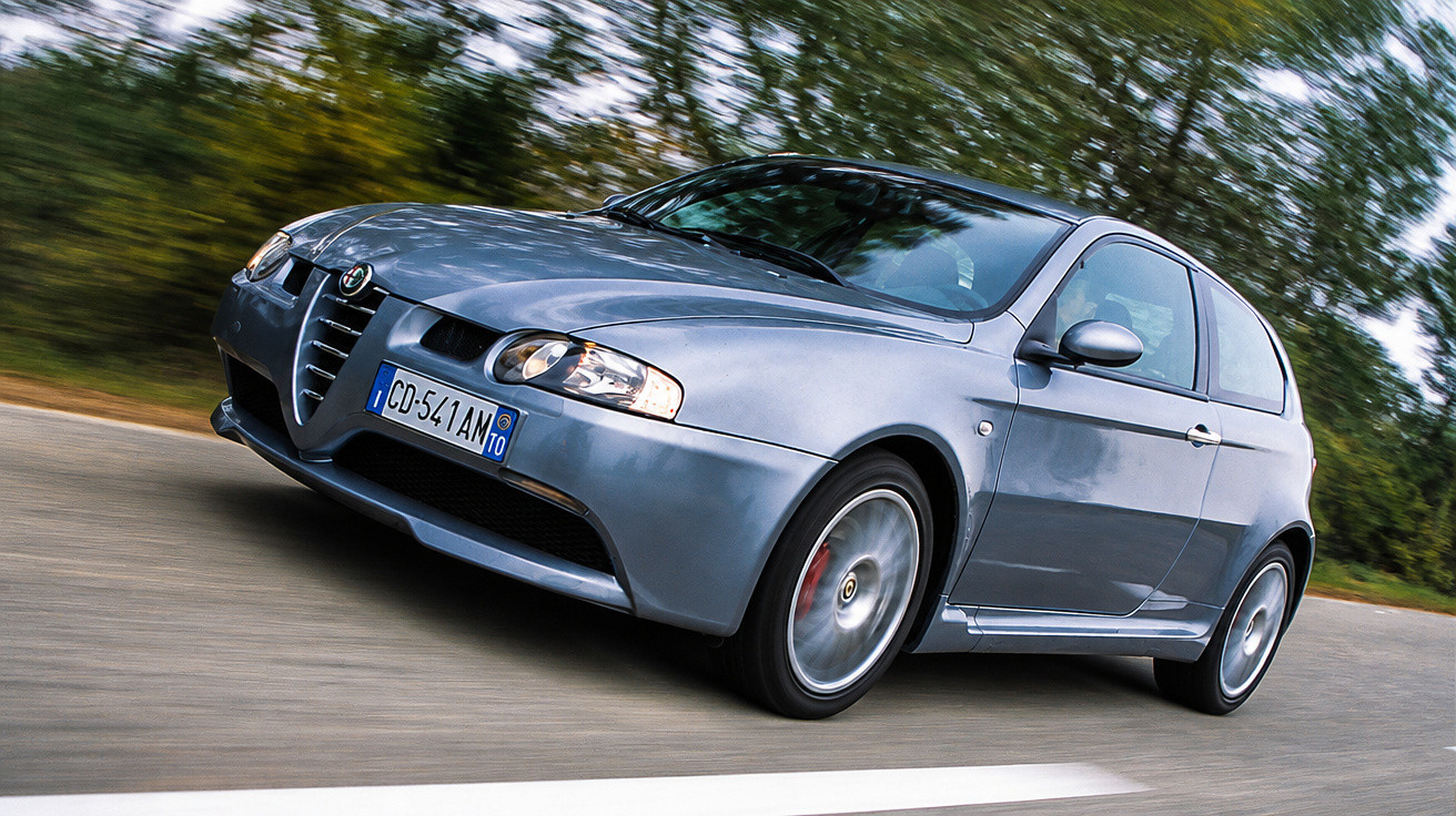 Carro cinzento Alfa Romeo a circular numa estrada com vegetação ao fundo.