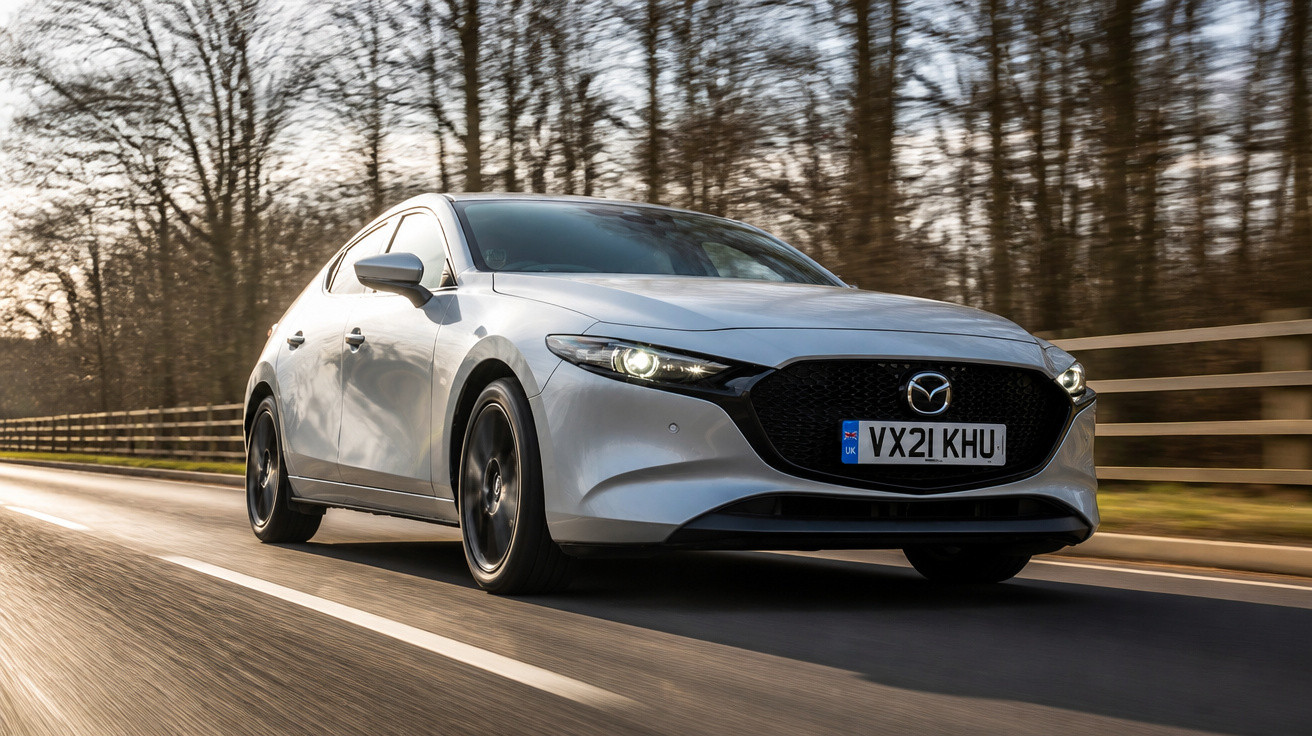 Carro Mazda cinzento a circular numa estrada com árvores sem folhas ao fundo em dia claro.