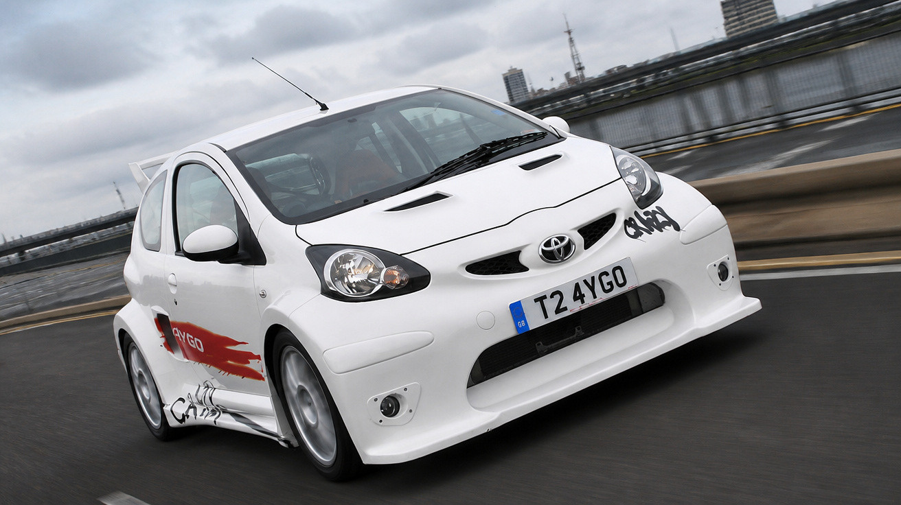 Carro desportivo branco Toyota Aygo em movimento numa estrada urbana junto a uma zona ribeirinha.