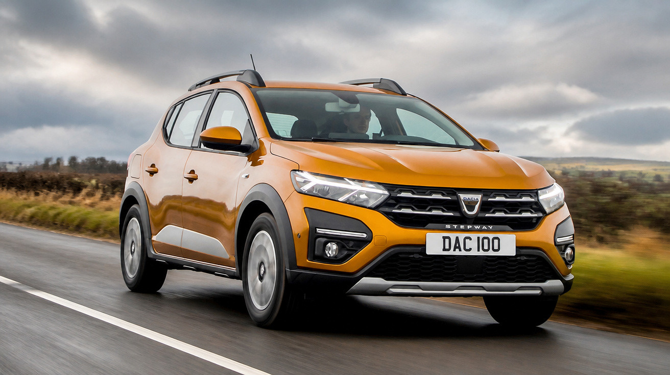 Carro Dacia Stepway laranja a circular numa estrada asfaltada com céu nublado.