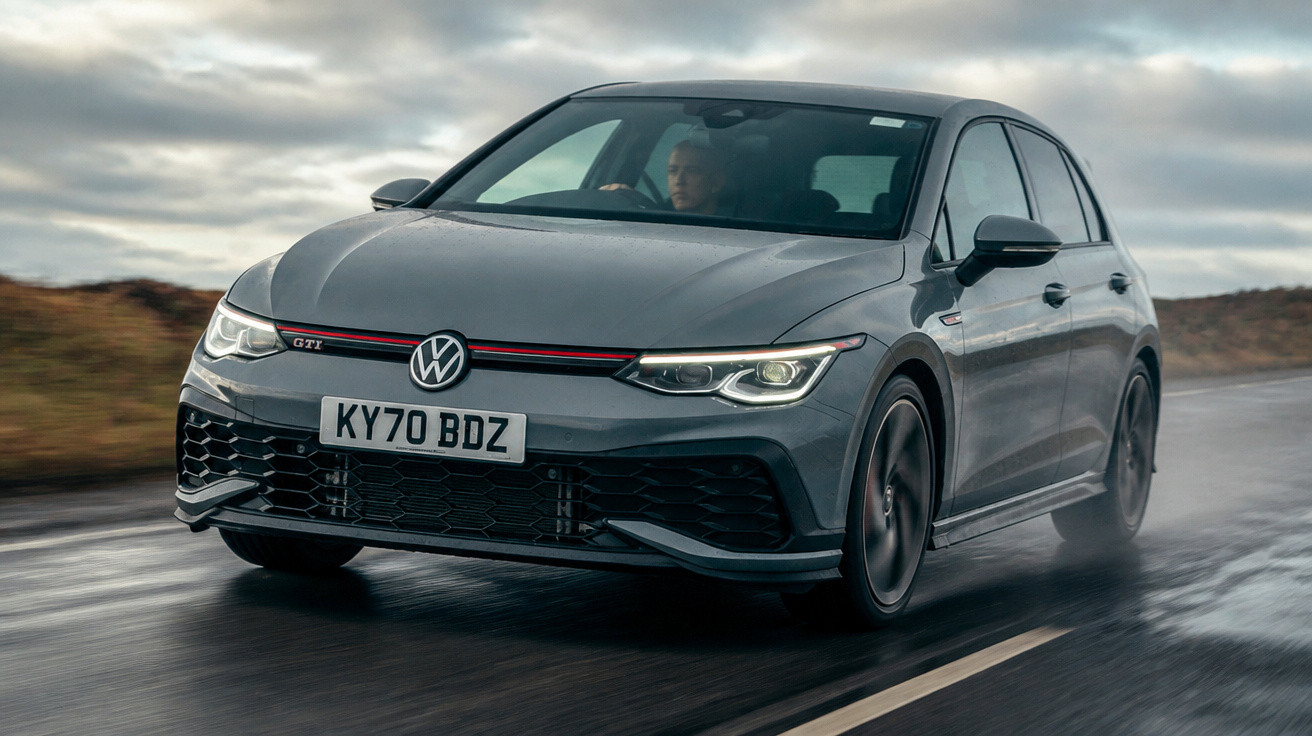 Carro Volkswagen Golf GTI cinza a conduzir numa estrada molhada sob céu nublado.