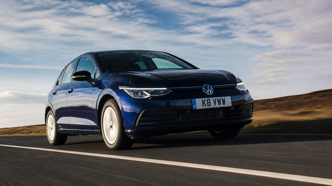 Carro Volkswagen Golf azul em estrada vazia com céu nublado ao fundo.