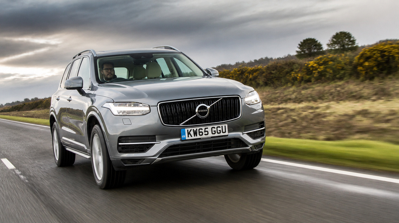Carro Volvo cinzento a circular numa estrada rodeada de vegetação sob céu nublado.