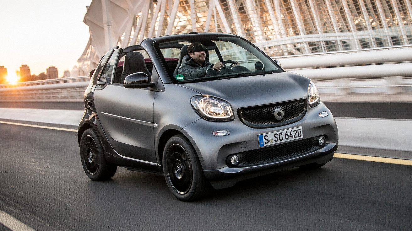Carro Smart Cabrio cinzento a circular numa estrada urbana com um condutor ao volante ao pôr do sol.