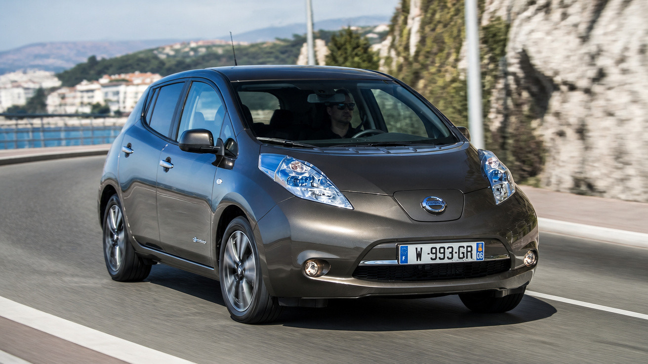 Carro elétrico Nissan Leaf cinzento a circular numa estrada costeira com paisagem montanhosa ao fundo.