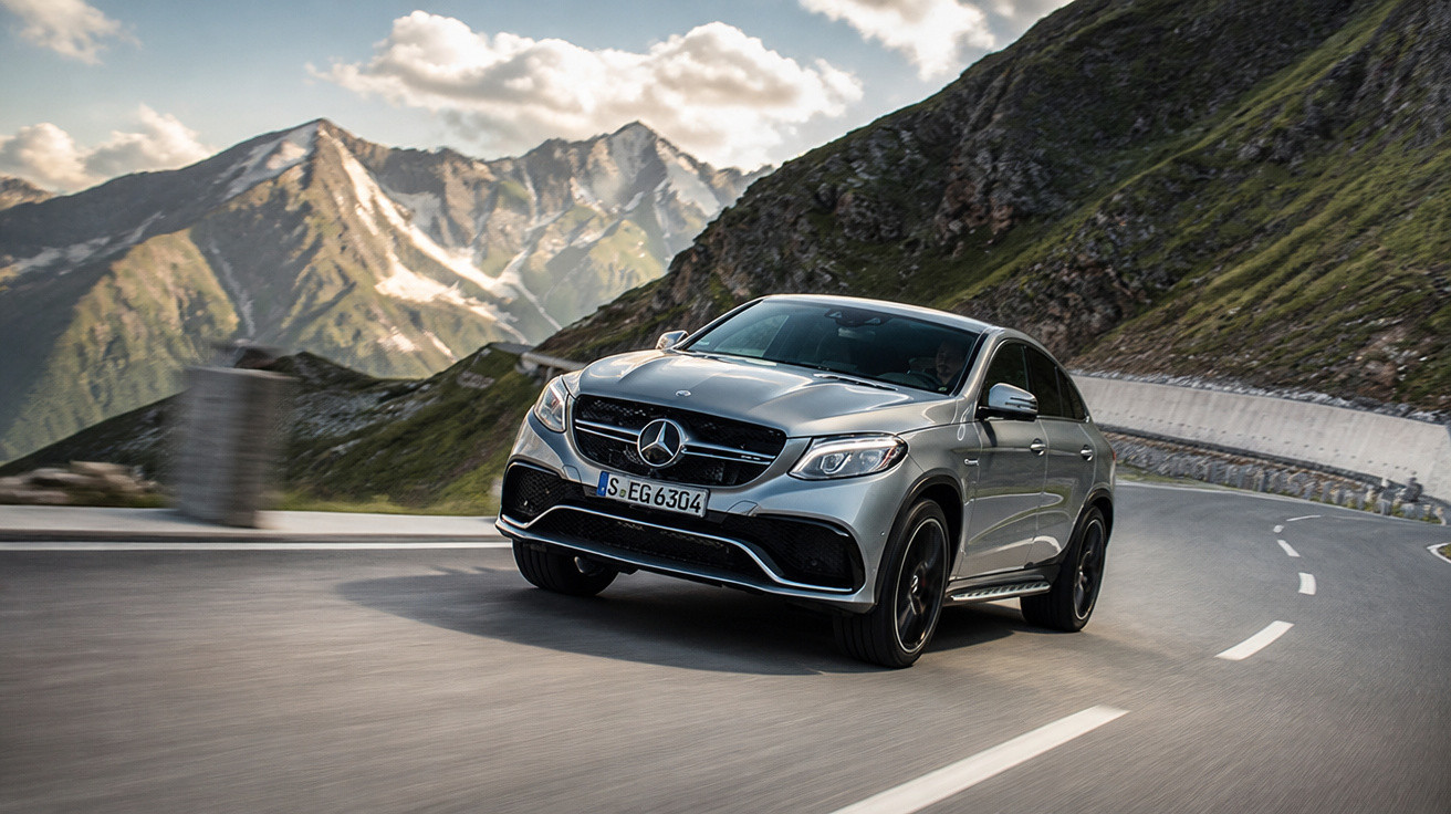 Carro Mercedes-Benz prata a conduzir numa estrada de montanha sinuosa com picos ao fundo.