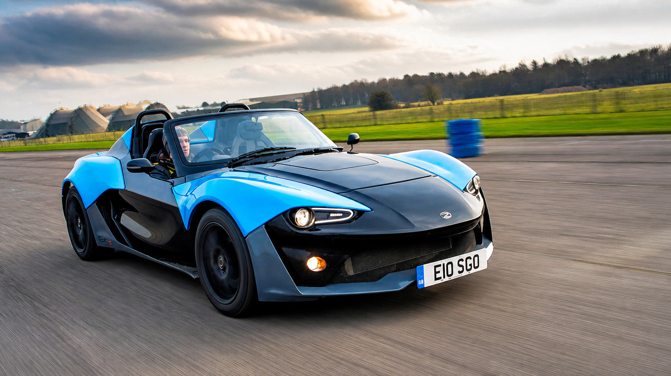 Carro desportivo azul e preto em velocidade numa pista de corrida com céu nublado ao fundo.