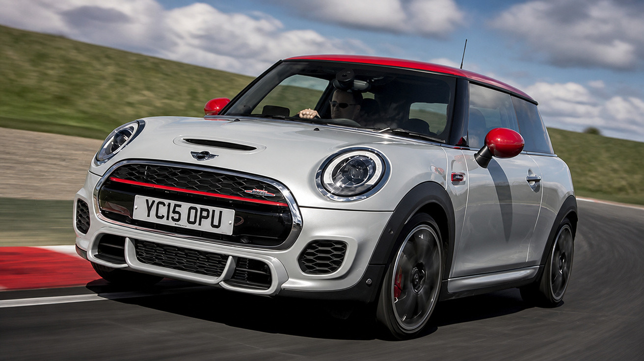 Mini Cooper branco e vermelho em velocidade numa pista de corrida sob céu nublado.