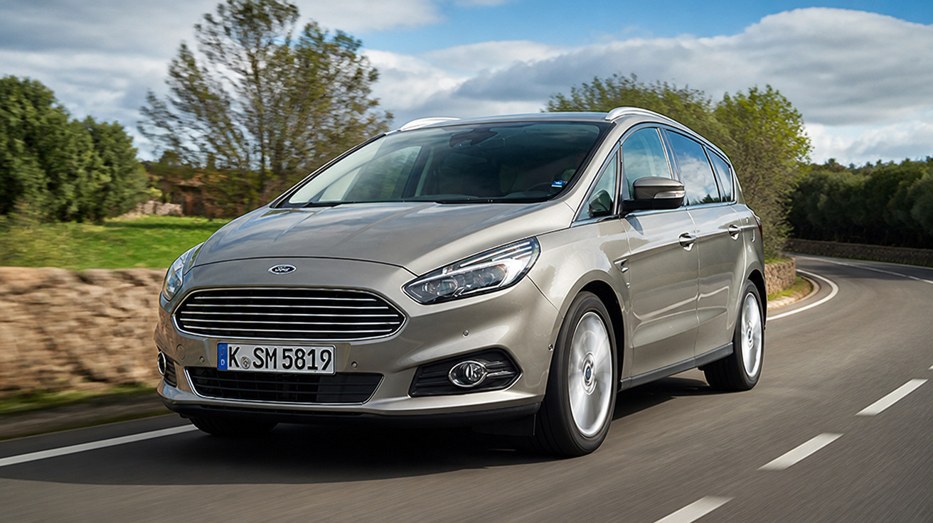Carro cinza prateado Ford em estrada curva com árvores e céu nublado ao fundo.