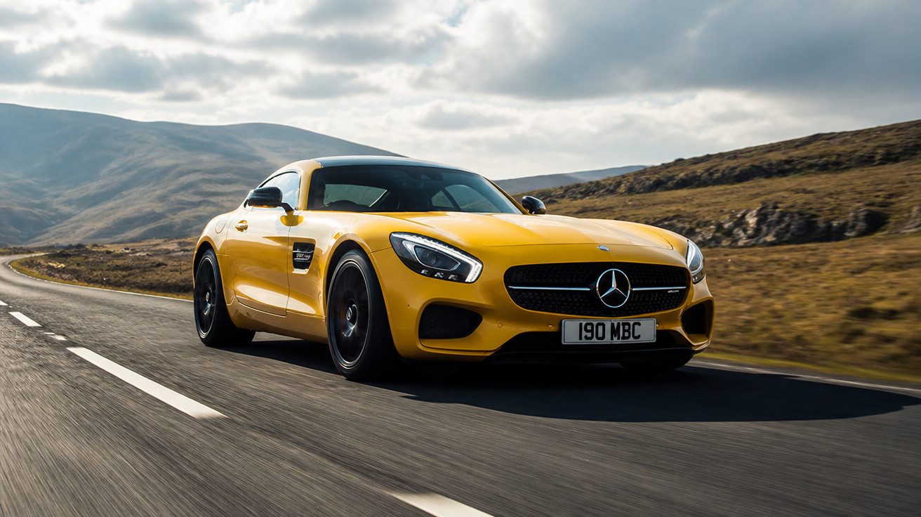 Carro desportivo amarelo Mercedes-Benz a circular numa estrada sinuosa no campo montanhoso sob céu nublado.