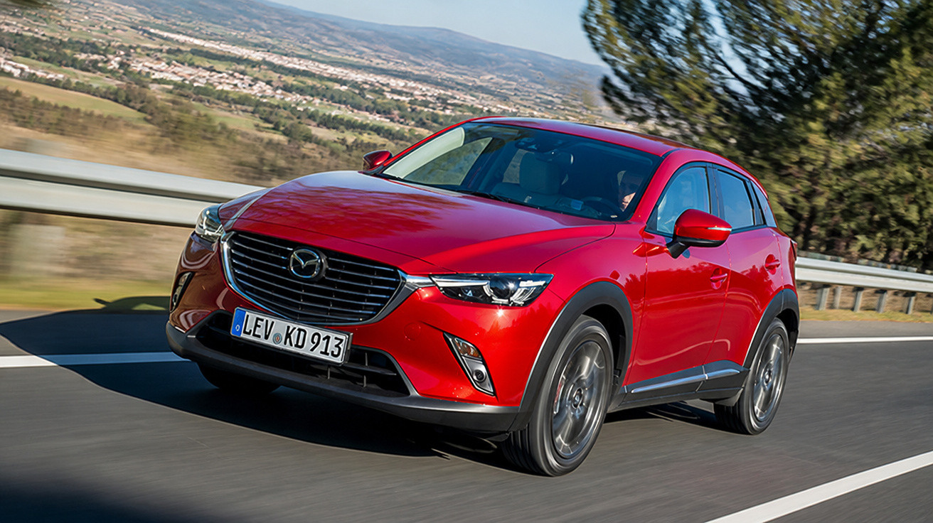Carro Mazda CX-3 vermelho em movimento numa estrada com paisagem e árvores ao fundo.